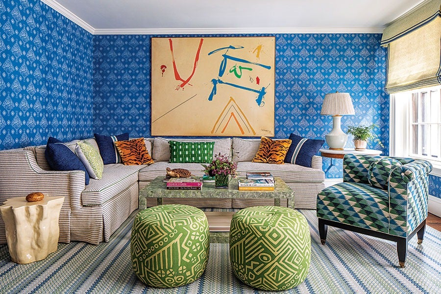 Living room with blue patterned wallpaper and a large abstract painting above a beige and white striped sectional sofa. The sofa has various throw pillows, including tiger print and green patterned ones. In front of the sofa is a glass coffee table with books and a flower arrangement. Two green patterned ottomans are placed in front of the coffee table. To the right, there is a blue and green geometric patterned armchair. A wooden side table with a white lamp and a small plant is next to the sofa by a window with beige Roman shades. The floor has a blue and white striped rug.