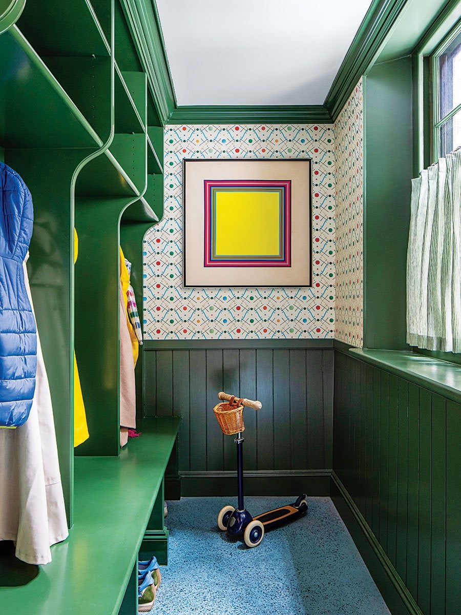 Mudroom with green built-in cubbies and bench, patterned wallpaper with colorful dots and lines, a framed geometric artwork featuring a yellow square, a blue scooter with a wicker basket, and a window with green trim and striped curtains.