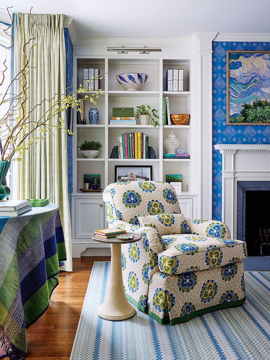 A cozy living room corner featuring a floral-patterned armchair with blue, green, and yellow designs. Next to the chair is a small round side table holding a few books and a dish. Behind the chair is a white built-in bookshelf filled with books, decorative bowls, plants, and framed art. The room has blue patterned wallpaper on one wall, a white fireplace mantel, and a blue and green striped rug on the wooden floor. A table covered with a green and blue plaid cloth and a vase with branches and yellow flowers is partially visible on the left. Cream-colored curtains frame a window beside the bookshelf.
