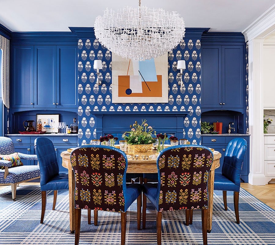 Dining room with a round wooden table surrounded by six chairs, four upholstered in blue leather and two with a dark fabric featuring colorful geometric floral patterns. The walls are painted deep blue with white floral wallpaper in the center section, flanked by blue cabinetry. A large, modern white chandelier hangs above the table. The floor has a blue and white plaid rug, and a modern abstract painting is centered on the wallpapered wall above a blue fireplace mantel.