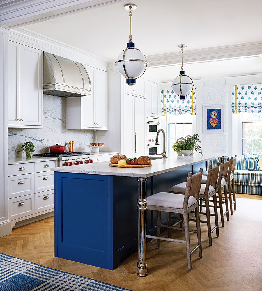 Modern kitchen with a large blue island featuring a white marble countertop and four wooden bar stools with light cushions. The surrounding cabinetry is white with brass handles, and a stainless steel range hood is mounted above a stove with red knobs. Two spherical pendant lights with brass and navy accents hang above the island. The floor is light wood in a herringbone pattern, and a blue patterned rug is partially visible. The background includes windows with white and blue patterned Roman shades and a striped blue and white cushioned bench.