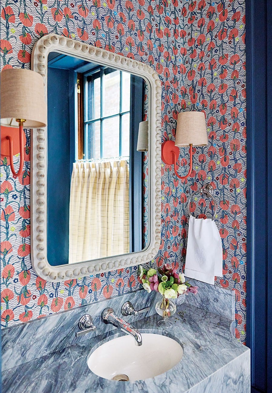Bathroom vanity with a gray marble countertop and an oval white sink. Above the sink is a large rectangular mirror with a decorative white frame. The walls are covered in a vibrant floral wallpaper featuring red flowers and blue stems. Two wall-mounted light fixtures with beige lampshades flank the mirror. A silver towel ring holds a white hand towel, and a small glass vase with a bouquet of flowers sits on the countertop. A window with blue trim and a beige curtain is reflected in the mirror.