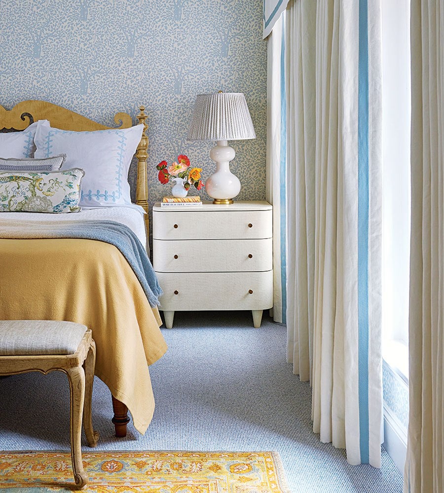 A cozy bedroom corner featuring a wooden bed with a mustard yellow blanket and light blue throw, adorned with white and patterned pillows. Next to the bed is a cream-colored three-drawer nightstand with a white ceramic lamp with a pleated shade and a small vase of colorful flowers. The wall behind has a light blue wallpaper with a subtle tree pattern. Cream curtains with blue vertical stripes frame a window, and a light blue carpet covers the floor, partially overlaid by a yellow and blue patterned rug. A wooden bench with a cushioned seat sits at the foot of the bed.