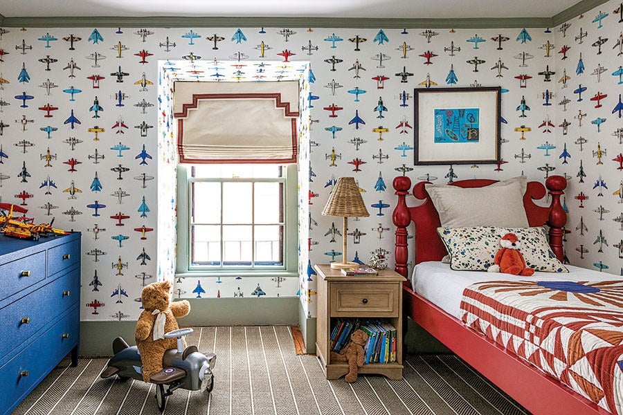 Children's bedroom with airplane-themed wallpaper featuring various colorful planes. The room has a red wooden bed with white and patterned pillows, a bedside table with a lamp and books, and a blue dresser. A teddy bear sits on a toy airplane on the floor, and another stuffed animal is on the bed. The window has a beige Roman shade with red trim. The carpet is striped in neutral tones.