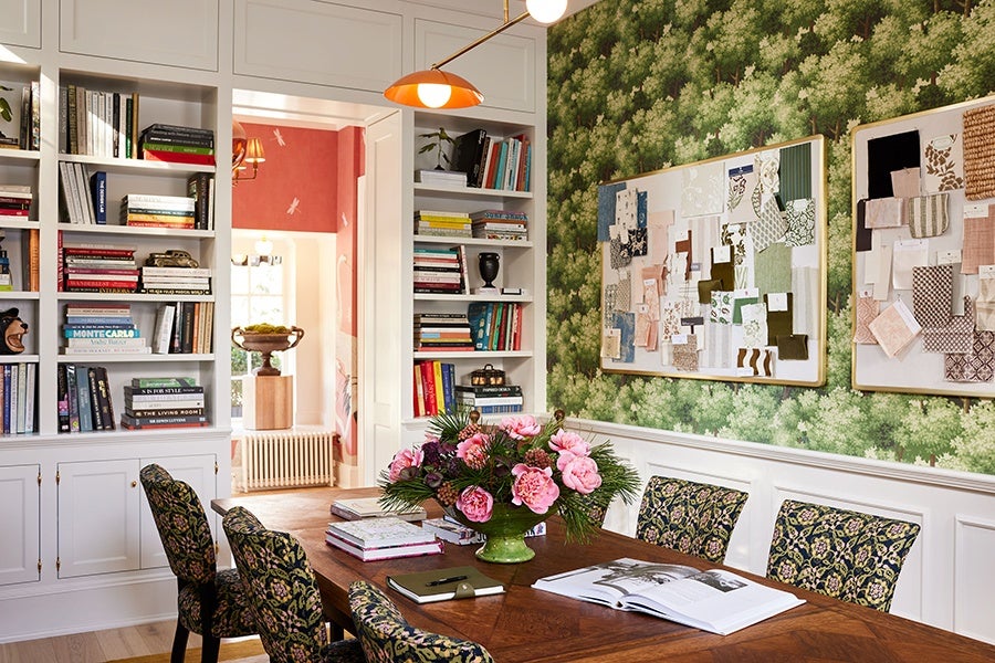 A cozy study or meeting room featuring a wooden table with a green vase holding a bouquet of pink flowers and greenery. The table is surrounded by chairs upholstered in a dark floral fabric. The walls are decorated with green leafy wallpaper and white wainscoting. Two large bulletin boards display various fabric swatches and papers. Built-in white bookshelves filled with books and decorative items flank a doorway leading to another room with red walls and a window. An orange pendant light hangs above the table.