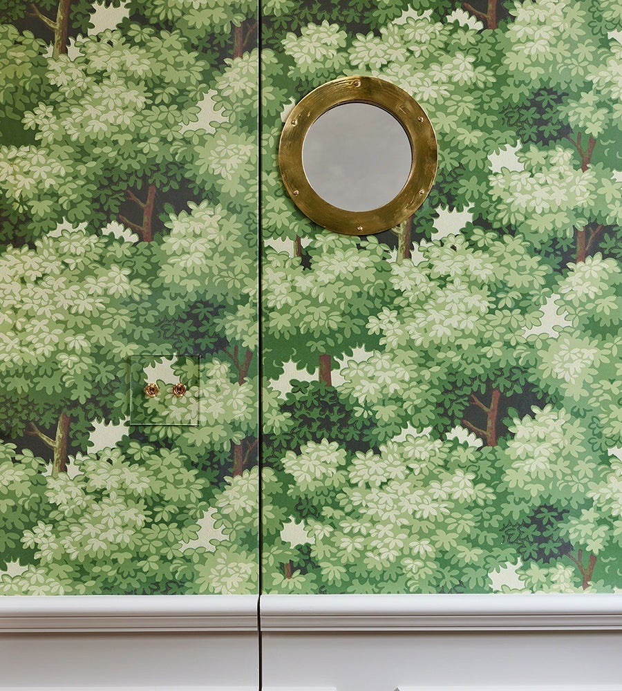 A wall covered with green leafy tree-patterned wallpaper featuring a small door with two round brass knobs and a round brass-framed porthole window above it. The bottom of the wall has white molding.