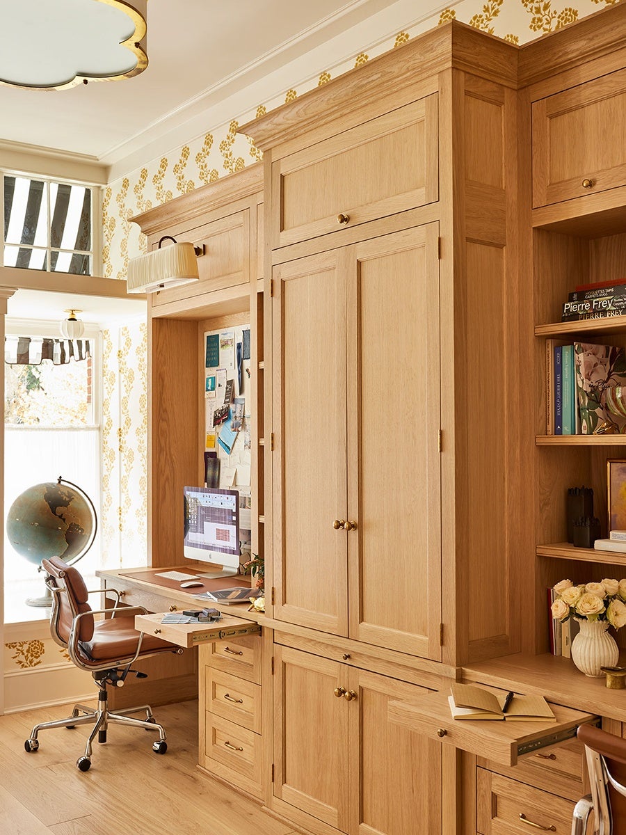 A built-in wooden home office setup with light oak cabinetry and drawers. The workspace includes a desk with a computer, a leather swivel chair, and a pull-out drawer with stationery. There is a bulletin board with pinned notes and photos above the desk. To the right, there are shelves holding books and a vase of flowers. The room has patterned wallpaper with a yellow floral design and a window with striped curtains, allowing natural light to enter. A globe is positioned near the window. The overall style is warm and classic with brass hardware on the cabinets.