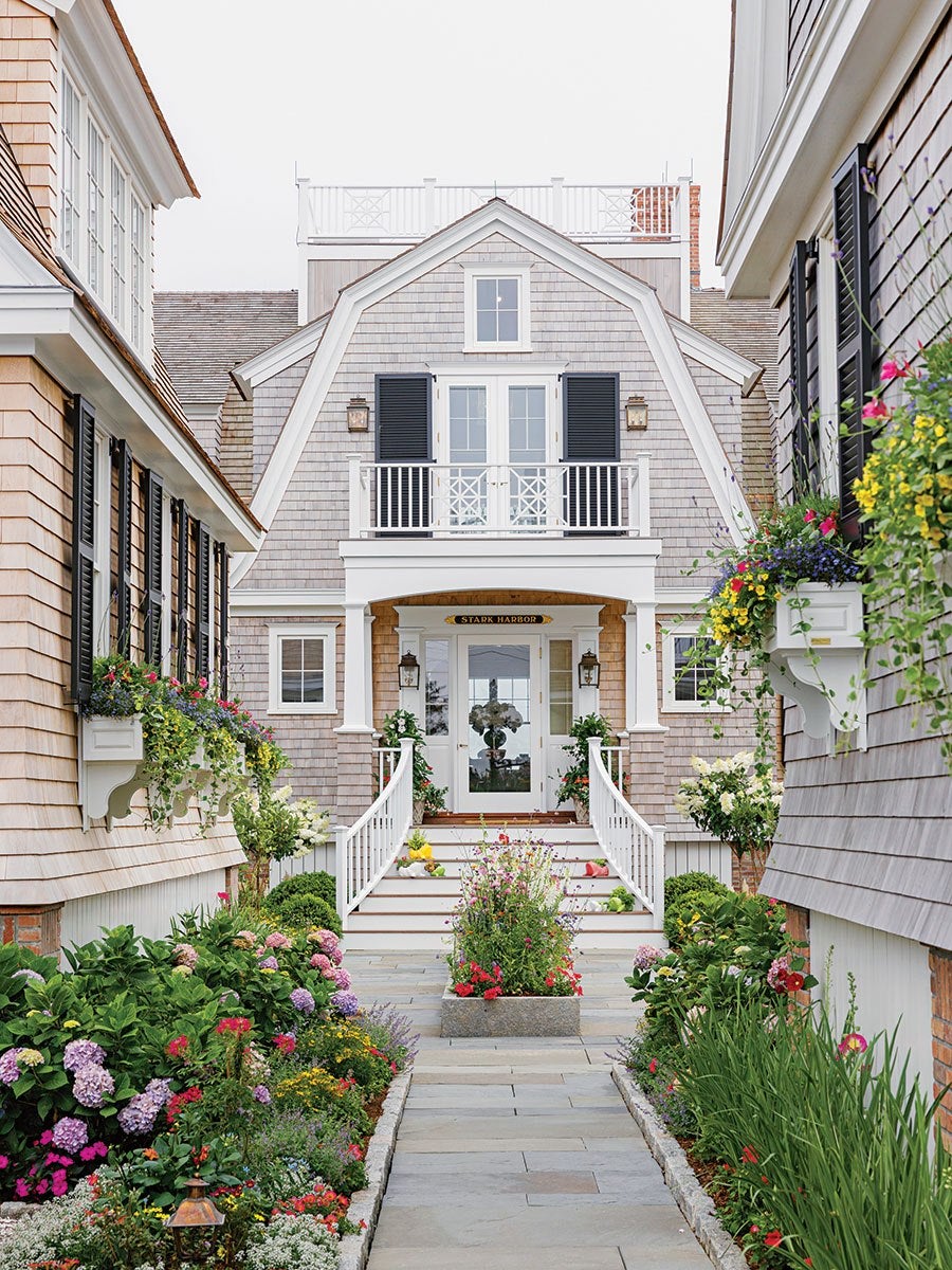 A charming house with gray shingle siding and white trim, featuring a central entrance with white double doors and a small balcony above with black shutters. The entrance is accessed by white stairs with railings, flanked by lush flower beds filled with colorful blooms and greenery. Window boxes with vibrant flowers adorn the windows on both sides of the walkway, creating a welcoming and picturesque scene.