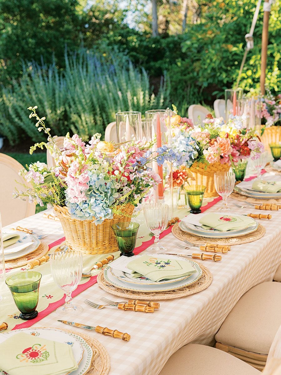 A long outdoor dining table set with a light beige and white checkered tablecloth and a floral table runner with a pink border. The table features wicker placemats, white plates with floral designs, and light green napkins embroidered with flowers. Bamboo-handled cutlery is neatly arranged beside each setting. Green and clear glassware, including goblets and water glasses, are placed at each setting. The centerpiece consists of multiple wicker baskets filled with pastel-colored flowers, including pink, blue, and white blooms, along with tall pink taper candles in clear glass holders. The setting is surrounded by beige cushioned chairs and lush green garden foliage in the background.