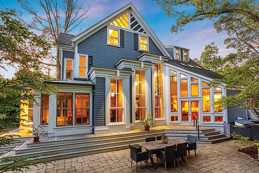 Large two-story house with blue siding and white trim, featuring numerous tall windows glowing warmly from interior lights. The house has a multi-level deck and a stone patio with a wooden table and six black chairs. Surrounding trees frame the scene under a colorful evening sky.