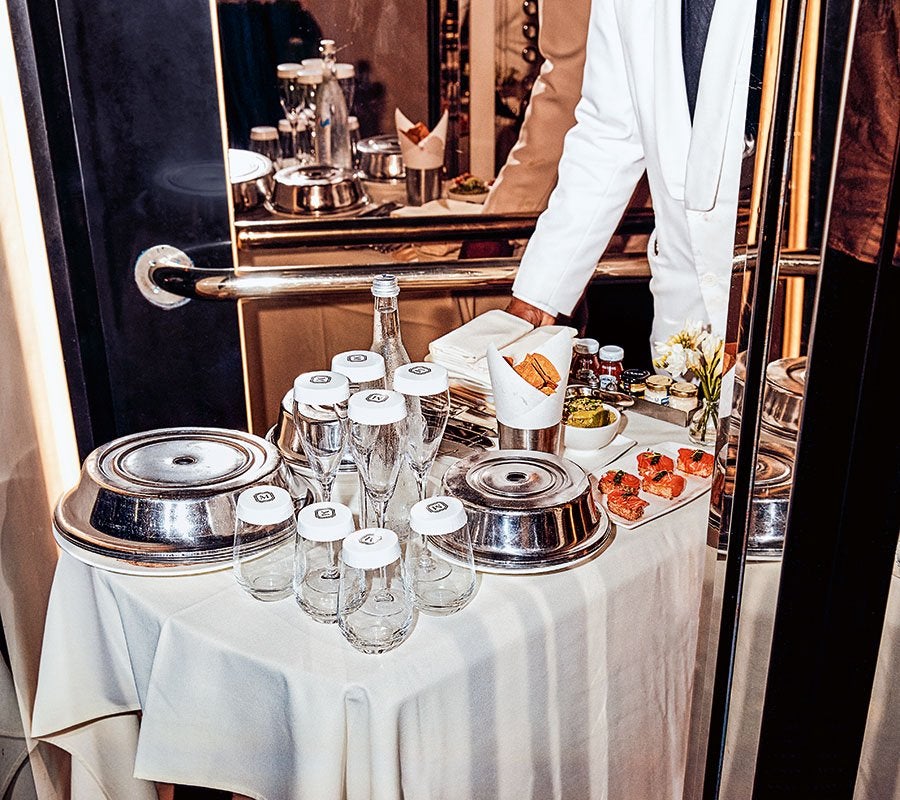 A table covered with a white tablecloth holds several upside-down glasses with white covers, two large covered silver serving dishes, a bottle of water, a small bowl of green dip, a plate with pieces of smoked salmon or similar fish garnished with herbs, a metal container with breadsticks wrapped in paper, a small vase with white flowers, and a stack of white napkins. A person wearing a white jacket is partially visible behind the table. The scene is reflected in a mirror behind the table.