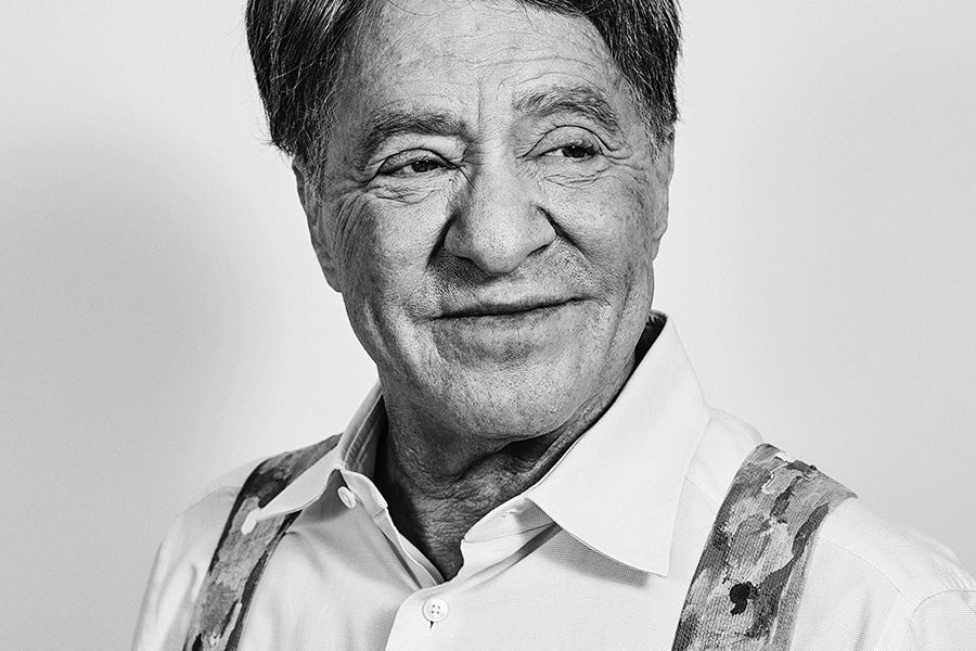 Black and white portrait of an older man with short dark hair, wearing a light-colored button-up shirt and patterned suspenders, looking slightly to the side with a gentle smile.