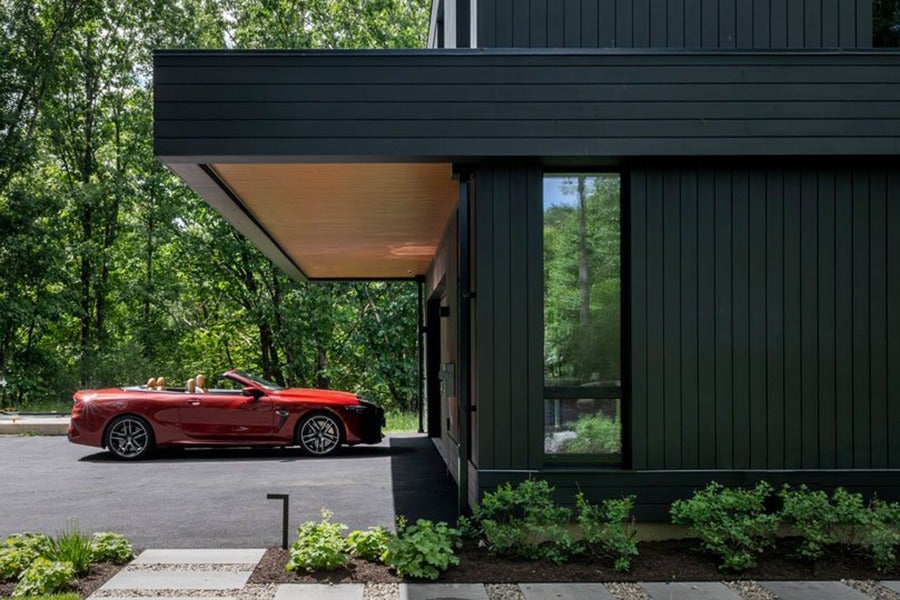 Red convertible sports car parked next to a modern black house with vertical siding, surrounded by greenery and trees. The driveway is paved, and there are small plants and stepping stones in the foreground.