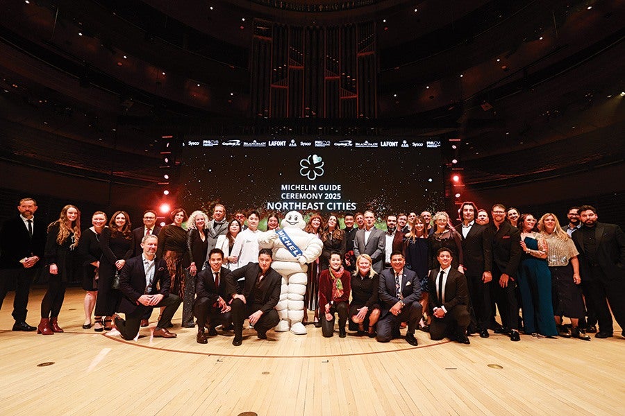 A large group of people dressed in formal and semi-formal attire posing on a wooden stage in front of a large screen that reads "Michelin Guide Ceremony 2025 Northeast Cities." The Michelin Man mascot stands in the center holding a blue sash with "MICHELIN" written on it. The background is dark with subtle lighting highlighting the group.