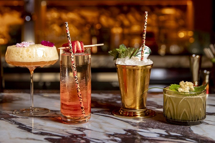 Four cocktails on a marble countertop: a frothy drink in a coupe glass garnished with pink flowers, a tall glass with a red gradient drink and strawberry slices on a skewer, a gold metal cup filled with crushed ice and mint leaves, and a short glass with a greenish drink garnished with herbs and a small flower. Each drink has a patterned straw.