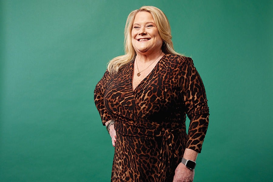 A smiling woman with blonde hair wearing a long-sleeved, leopard-print dress. She has a necklace, bracelets, and a smartwatch on her left wrist, standing against a solid green background.