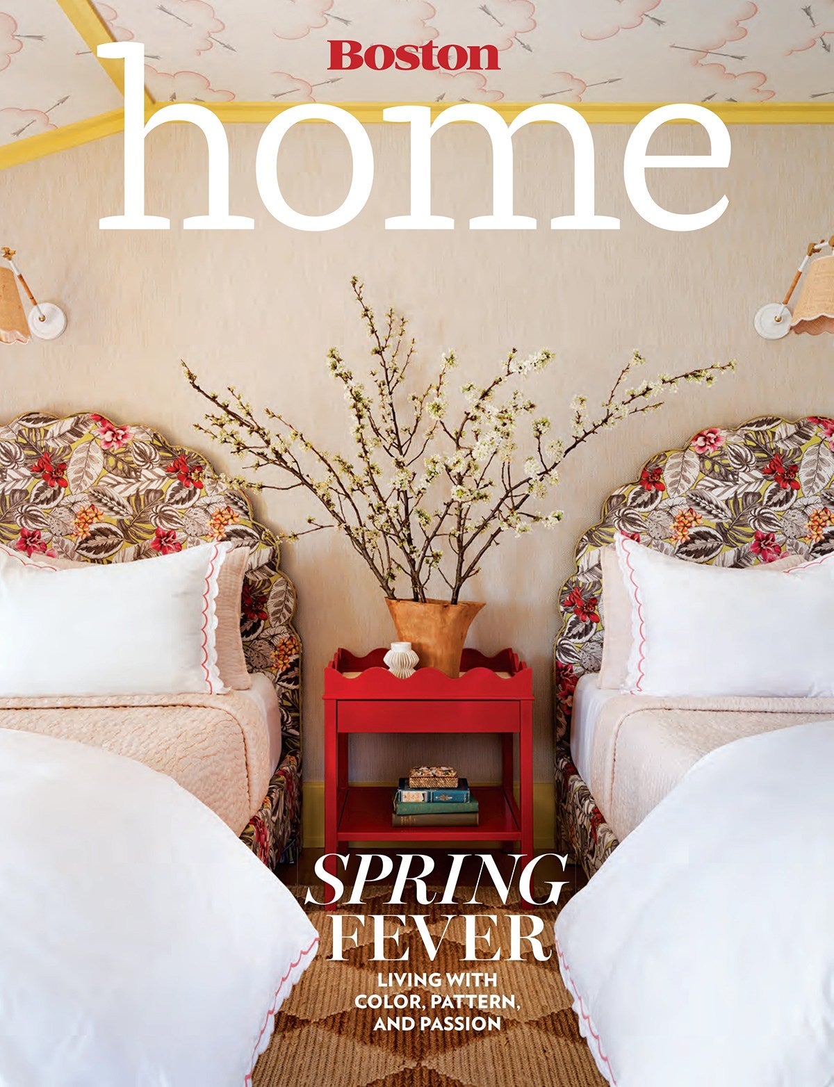 Two twin beds with floral-patterned headboards and white bedding featuring pink scalloped edges flank a bright red nightstand. On the nightstand is a terracotta vase filled with blossoming branches, a small white decorative object, and a few stacked books. The walls are a soft beige, and the ceiling has a light pattern with yellow trim. The floor has a textured, natural fiber rug. The overall style is colorful and inviting, emphasizing spring themes.