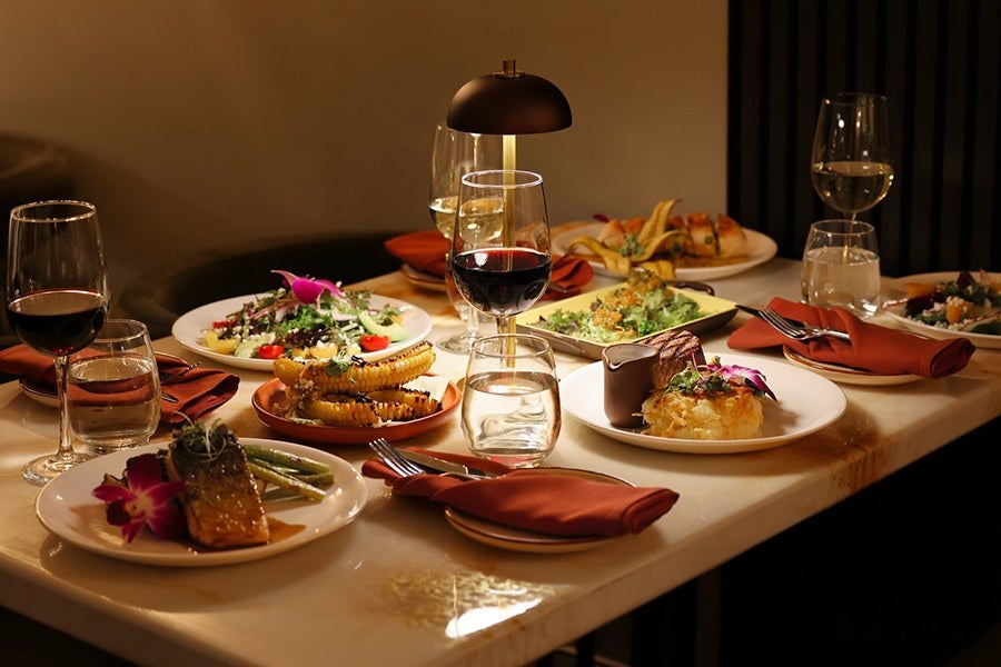 A dining table set with various plated dishes including grilled corn, a salad with cherry tomatoes and greens, a piece of cooked fish with green beans and an edible flower garnish, a dessert with chocolate and a flower garnish, and a plate of fries. The table also has several glasses of red and white wine, water glasses, and rust-colored cloth napkins with forks. A small, modern table lamp provides warm lighting.