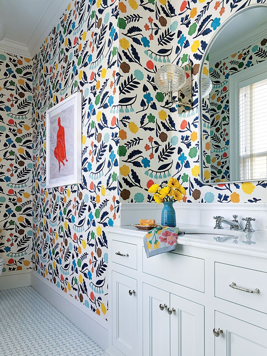 Bathroom corner with white cabinetry and a marble countertop. The walls are covered in colorful floral wallpaper featuring leaves and flowers in blue, green, yellow, orange, and brown. A large arched mirror hangs above the sink, reflecting a window with white blinds. A blue vase with yellow flowers and a soap dish with a bar of soap sit on the countertop, along with a floral-patterned hand towel. A framed artwork with a red figure is mounted on the adjacent wall. The floor has a white tile pattern.