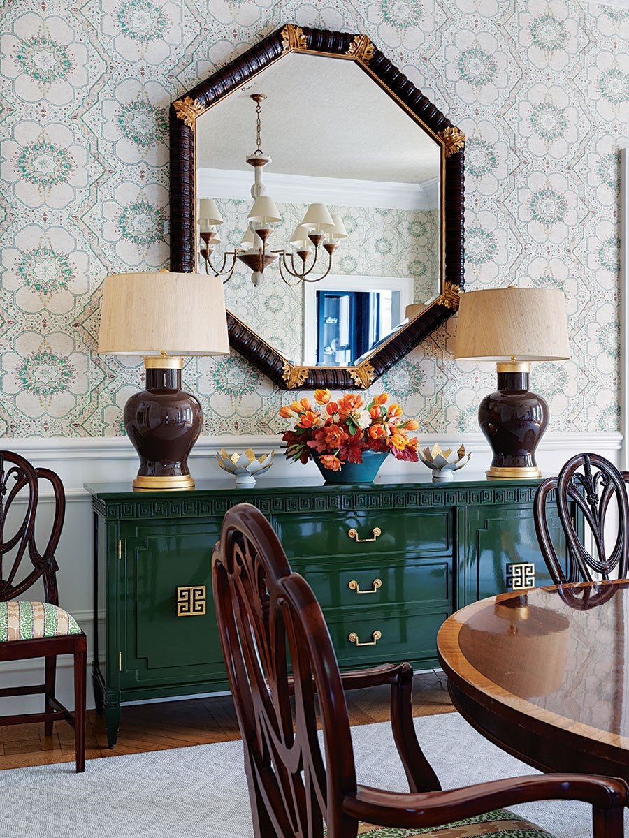 A dining room features a dark green sideboard with gold hardware, topped with two dark brown lamps with beige shades and a floral arrangement in a blue vase. Above the sideboard hangs a large octagonal mirror with a dark frame accented by gold details. The wallpaper has a vintage floral and geometric pattern in soft green and beige tones. Dark wooden chairs with intricate backs surround a polished wooden dining table. The room has a classic, elegant style with a mix of dark wood and vibrant green.