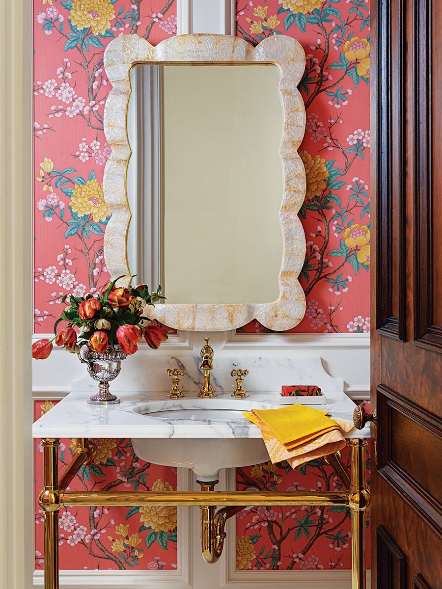 A bathroom vanity with a white marble countertop and an under-mount sink, supported by gold metal legs and fixtures. Above the sink is a large, scalloped-edge mirror with a textured, light-colored frame. The wall behind features vibrant pink floral wallpaper with yellow and green accents. On the countertop, there is a silver vase holding red and orange tulips, a small soap dish with a bar of soap, and two folded yellow and orange hand towels. A partially open dark wooden door is visible on the right side.
