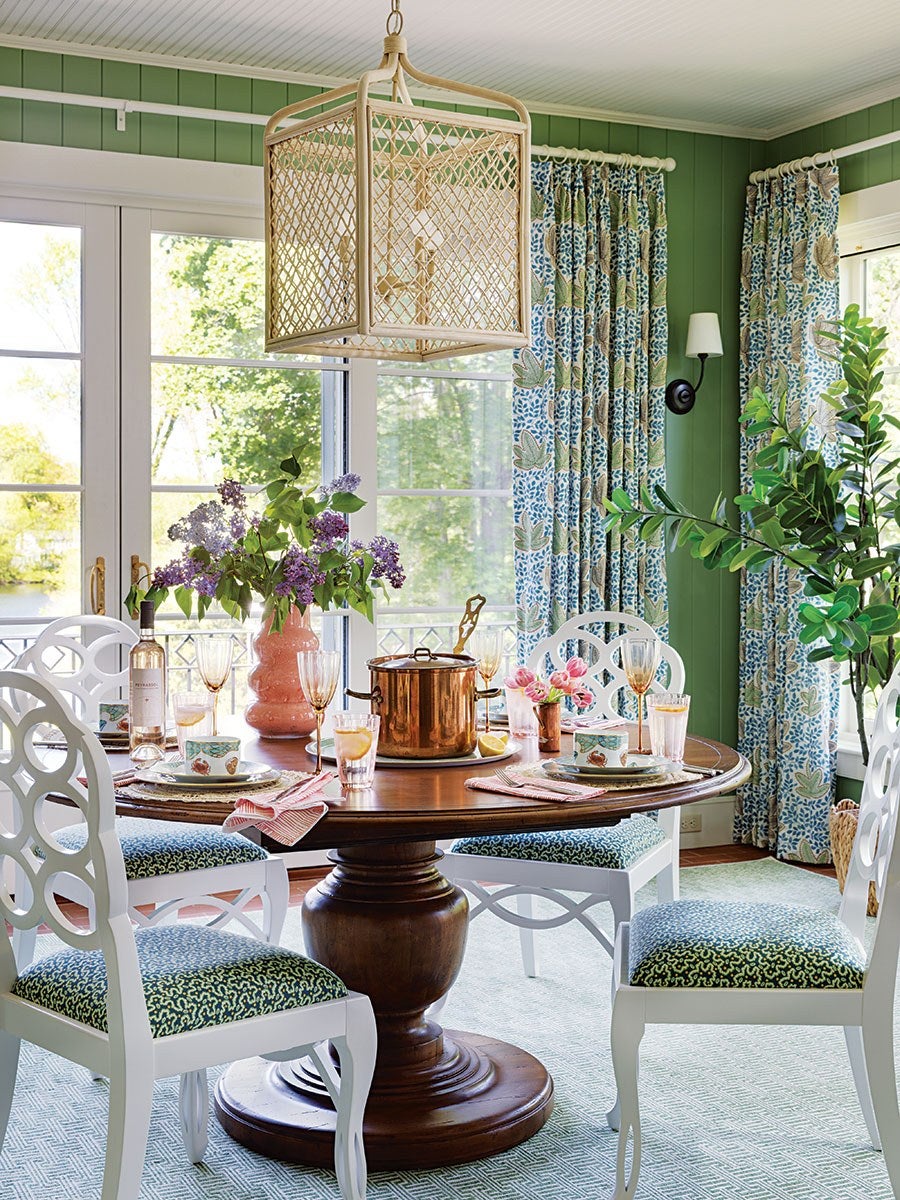 Round wooden dining table with a large central pedestal base, surrounded by white chairs with intricate circular back designs and patterned blue-green cushions. The table is set with glassware, plates, a copper pot, a bottle of wine, and a pink vase holding purple flowers. The room features green paneled walls, large windows with floral blue and green curtains, a woven pendant light fixture, and a leafy green plant in the corner.