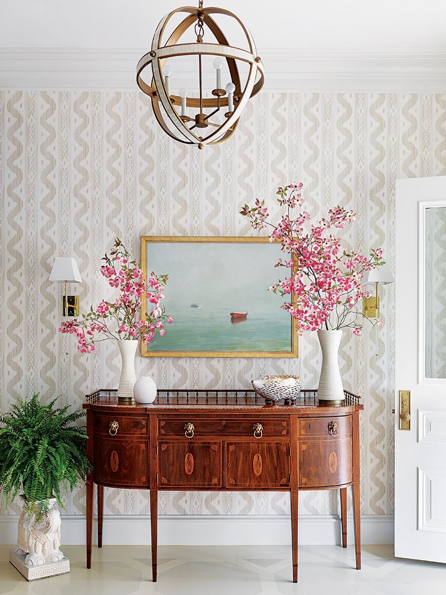 A wooden sideboard with intricate inlay details stands against a patterned beige and white wallpapered wall. On top of the sideboard are two white vases filled with pink flowering branches, a white decorative egg, and a white and gold bowl. Above the sideboard hangs a framed painting of a misty seascape with two small boats. To the left of the sideboard is a green fern plant in a white ornate planter. A white door with a brass handle is partially open on the right. A spherical wooden chandelier with exposed bulbs hangs from the ceiling. Two wall sconces with white shades flank the painting.