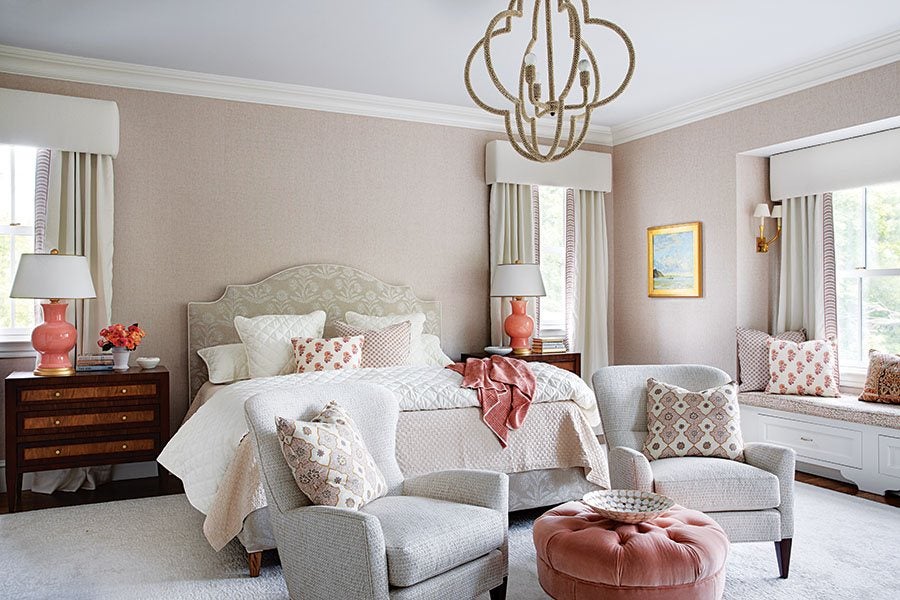 Bedroom with a beige upholstered headboard bed dressed in white and light pink bedding, flanked by dark wooden nightstands with coral-colored lamps. Two light gray armchairs with patterned pillows face a round, tufted pink ottoman. A window seat with cushions is set into a bay window on the right, and a rope chandelier hangs from the ceiling. The walls are light beige with white crown molding, and a small framed painting is on the right wall.