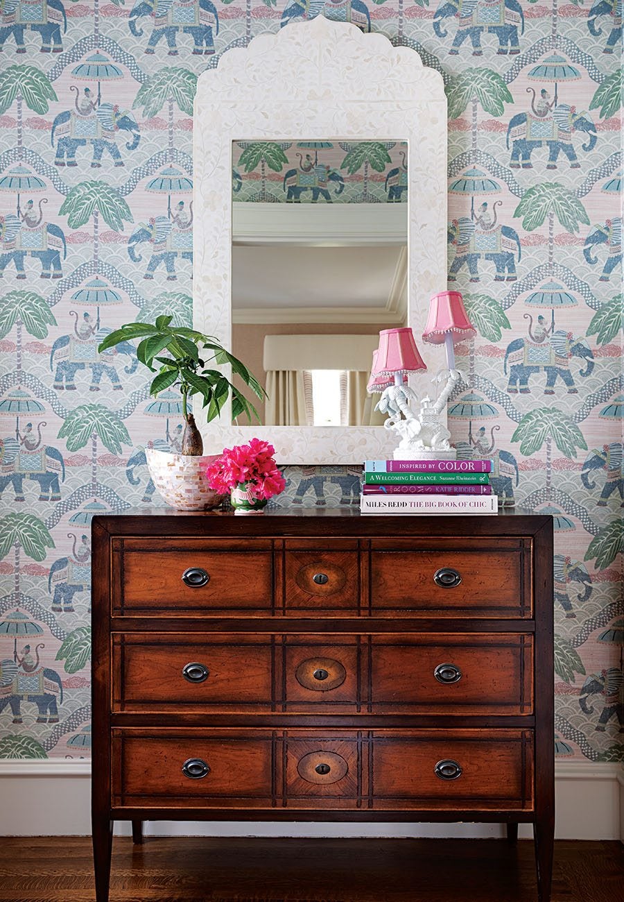 Dark wooden dresser with three drawers, topped with a white decorative mirror, a potted plant in a mosaic-style pot, a small vase of bright pink flowers, a stack of colorful books, and a white lamp with two pink lampshades. The background features wallpaper with a pattern of blue elephants, green palm trees, and monkeys under umbrellas.