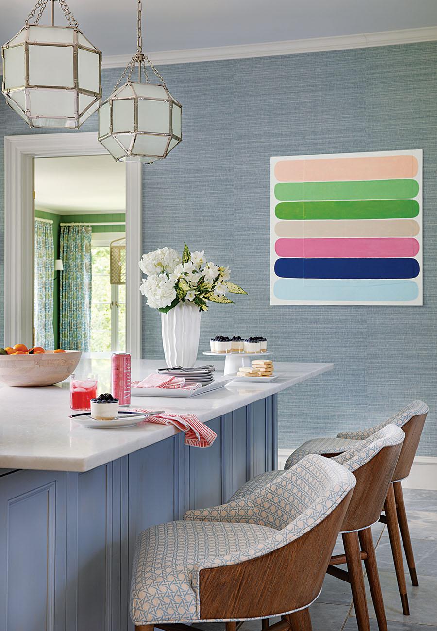 Kitchen island with a white marble countertop and blue paneled base, featuring three wooden bar stools with patterned upholstery. On the countertop, there is a white vase with white flowers, a wooden bowl with oranges, a pink drink in a glass, a pink can, plates with desserts, and red-striped napkins. Two geometric pendant lights hang above the island. The background wall is covered in textured blue wallpaper with a colorful abstract painting featuring horizontal rounded bars in pastel shades of peach, green, beige, pink, navy blue, and light blue. A doorway reveals a room with green walls and floral curtains.