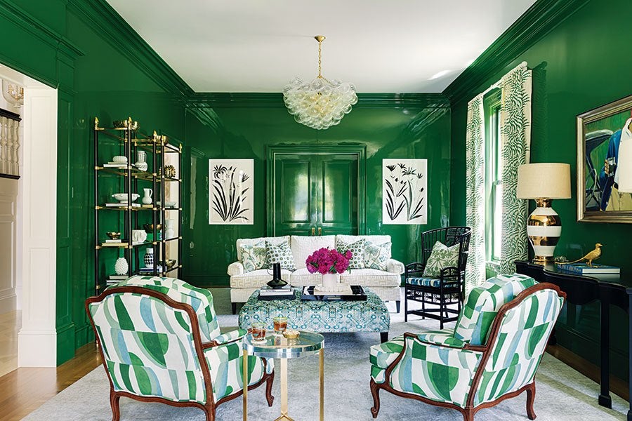 A living room with glossy dark green walls and trim, featuring a white sofa with patterned green and white pillows, two armchairs with green and white abstract upholstery, and a blue patterned ottoman. The room includes a gold and glass shelving unit, a black side table with a striped gold and white lamp, botanical curtains, black and white botanical artwork, and a chandelier with a cluster of glass orbs. A small round side table with drinks is placed between the armchairs.