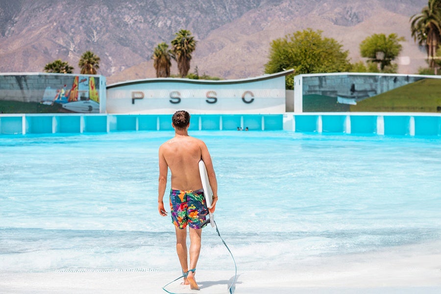 A shirtless man wearing colorful floral swim trunks is walking toward a large, empty wave pool while holding a white surfboard. The pool has clear blue water and is surrounded by a low wall with the letters "PSSC" displayed. In the background, there are palm trees and mountains under a clear sky.