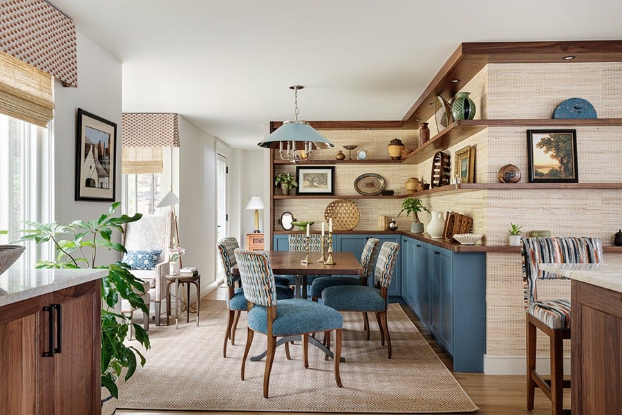 Dining area with a wooden table surrounded by six upholstered chairs featuring blue and patterned fabric. Blue cabinetry lines the back wall, topped with wooden shelves displaying decorative items and framed artwork. A light blue pendant lamp hangs above the table. The space is accented with a beige textured rug, a large leafy plant, and natural light from windows on the left. The walls have a light, woven texture, and there is a striped upholstered bar stool on the right.