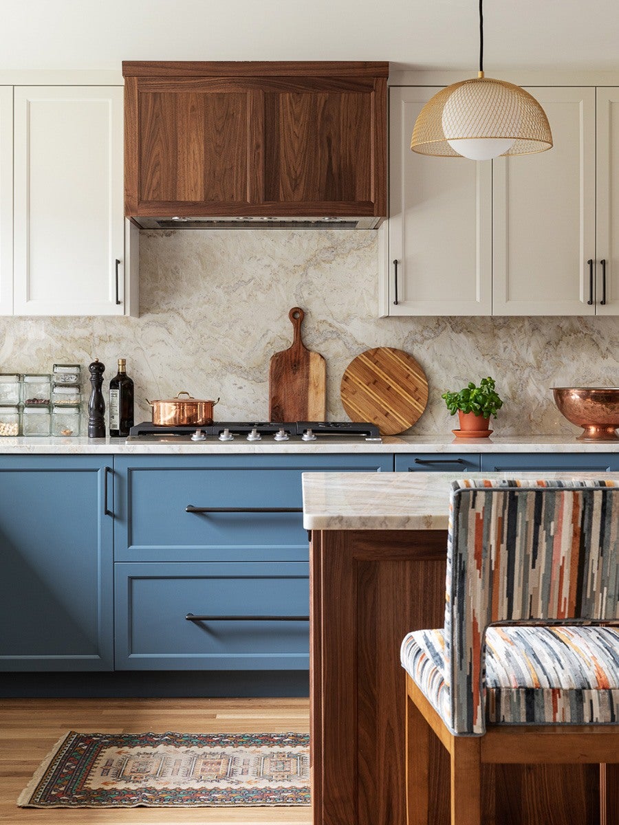 Kitchen with blue lower cabinets and white upper cabinets, featuring a wooden range hood. Marble backsplash and countertop with a gas stove. Wooden cutting boards, a copper pot, a small potted plant, and a copper bowl on the counter. A patterned upholstered chair and a small rug on the wooden floor. A woven pendant light hangs above.