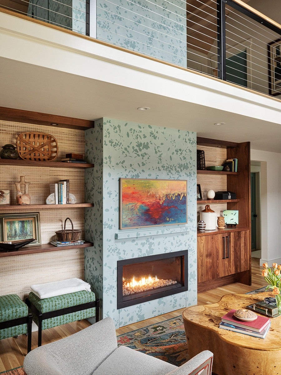 A modern living room features a light blue fireplace with a subtle leafy pattern, a rectangular gas firebox with white stones, and an abstract colorful painting above it. To the left, wooden shelves hold books, baskets, and decorative items against a textured beige wall. To the right, a wooden cabinet with shelves displays more decor. In the foreground, a natural wood coffee table with books and a vase of orange tulips sits on a patterned rug, and a light gray upholstered chair is partially visible. A metal railing with horizontal cables lines the upper level.