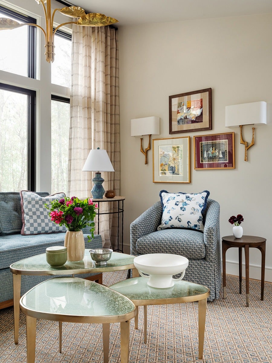A cozy living room corner featuring a blue patterned armchair with a white and blue floral pillow, a blue sofa with checkered and solid pillows, and three nested glass-top coffee tables with gold legs. On the tables are a beige vase with pink and purple flowers, a green ceramic container, a small silver bowl, and a white decorative bowl. A small wooden side table holds a white vase with dark red flowers. The wall behind has three framed artworks and two gold and white wall sconces. Light beige curtains hang beside large windows, and a blue ceramic table lamp sits on a round side table next to the sofa. The floor is covered with a patterned carpet.