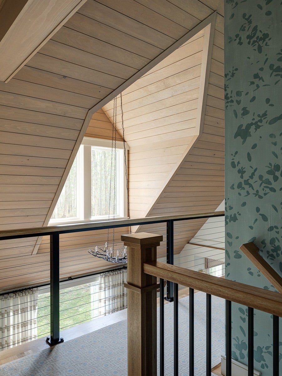 A modern interior space featuring a wooden staircase railing with black metal balusters. The ceiling and walls are covered in light wood paneling with an angular, geometric design. A large window lets in natural light, and a unique chandelier with a branch-like design hangs from the ceiling. The adjacent wall is painted teal with a subtle floral pattern. Light-colored curtains hang below the window, and a light blue carpet covers the floor.