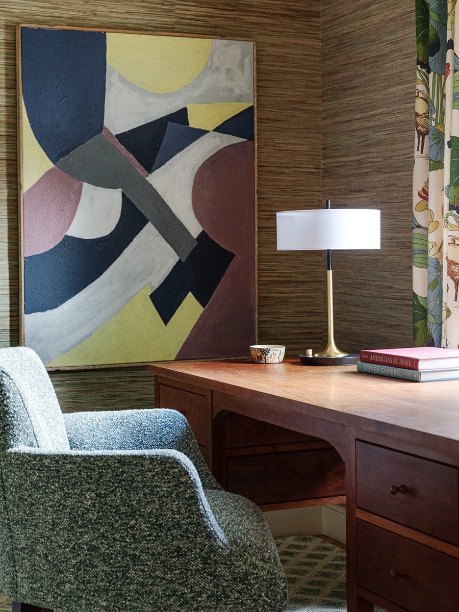 Mid-century style wooden desk with drawers, topped with a modern table lamp with a white shade, a small decorative bowl, and two stacked books. A textured blue-gray upholstered chair is positioned in front of the desk. The background features a large abstract painting with geometric shapes in muted colors and grasscloth wallpaper. A floral patterned curtain is partially visible on the right side.