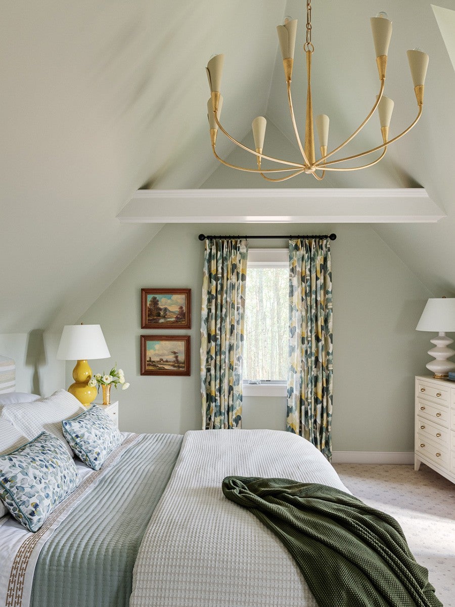 Bedroom with a sloped ceiling painted in light gray-green, featuring a bed with white and light blue bedding, floral-patterned pillows, and a dark green throw blanket. The window is framed by curtains with a blue, green, and yellow abstract pattern. Two framed landscape paintings hang on the wall beside the window. There are yellow and white bedside lamps on white nightstands, and a white dresser with a white lamp on the right side. A modern gold chandelier with cream-colored shades hangs from the ceiling.