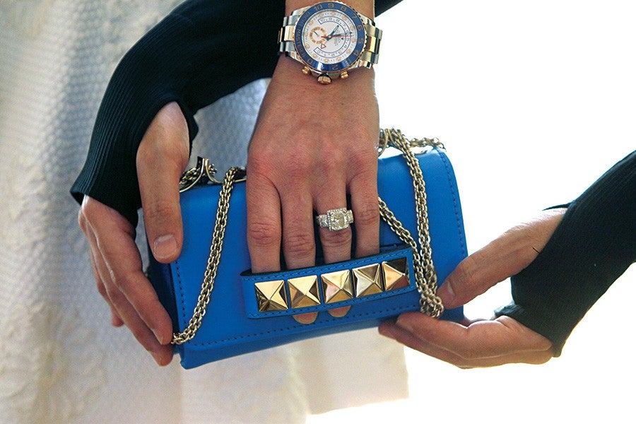 A person wearing a silver and gold watch and a large diamond ring is holding a small blue handbag with a gold chain strap and gold pyramid studs on the front. The person is dressed in black sleeves with thumb holes.