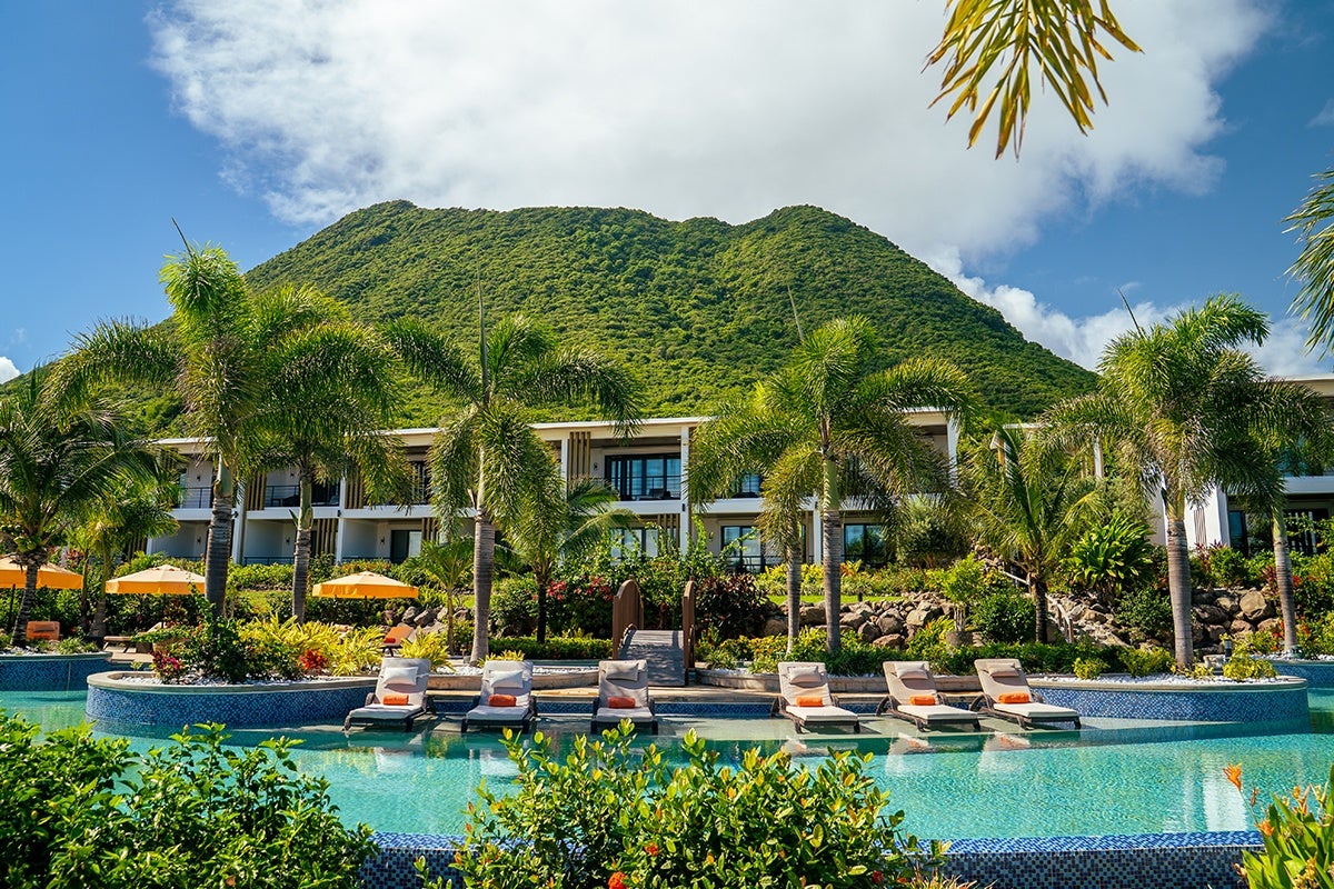 Resort pool area with multiple lounge chairs partially submerged in the water, surrounded by palm trees and lush greenery. Behind the pool is a modern two-story building with balconies, set against a backdrop of a large green mountain under a partly cloudy sky.