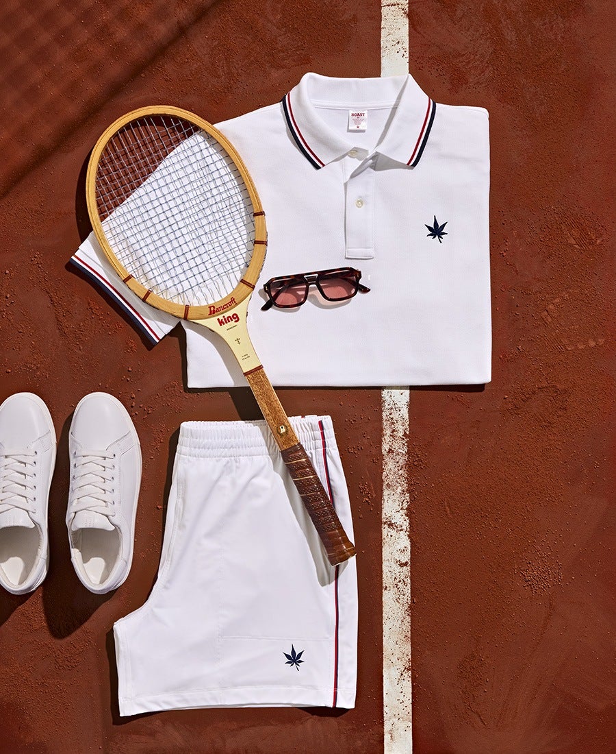 White tennis outfit consisting of a folded polo shirt and shorts, both featuring a small navy blue leaf emblem. The polo has a collar with red and navy stripes. A vintage wooden tennis racket with a brown grip and the brand name "Dancraft king" in red is placed diagonally across the outfit. A pair of white sneakers and dark sunglasses with a brown frame are also included, all arranged on a red clay tennis court with a white line running vertically.