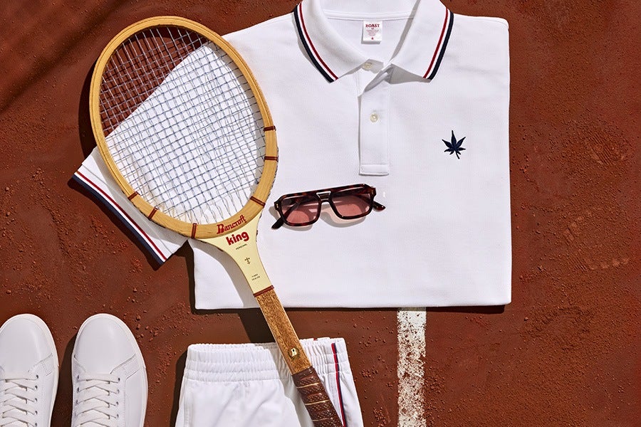 White polo shirt with red and black striped collar and a small dark leaf emblem on the chest, paired with white shorts and white sneakers. A vintage wooden tennis racket with "Bancroft king" written on it and a pair of tortoiseshell sunglasses with pink lenses are also included, all arranged on a clay tennis court.
