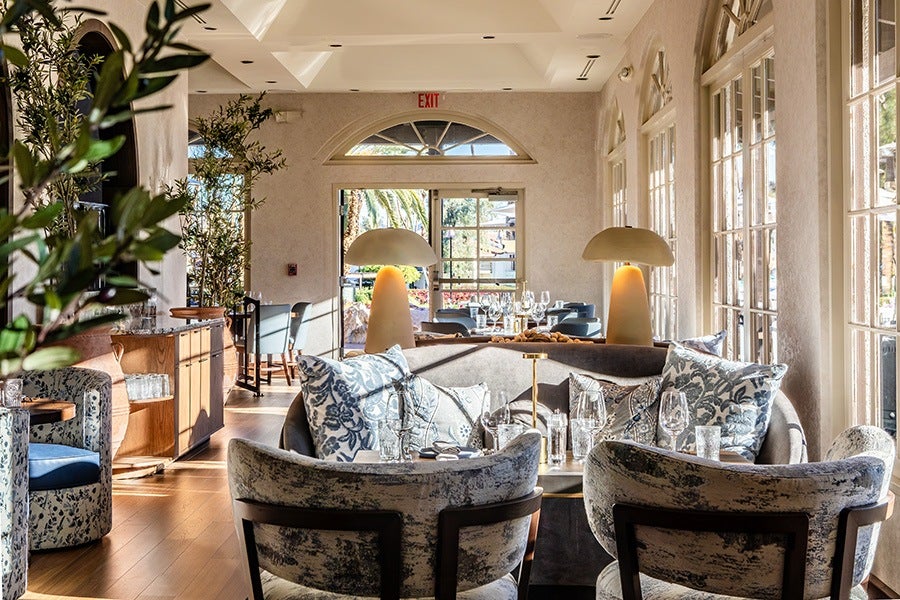 A bright, elegant dining area with large windows letting in natural light. The space features a curved sofa with patterned cushions, two matching armchairs, and a wooden table set with glassware and a small centerpiece. Two modern table lamps with beige shades are positioned behind the sofa. The room has light-colored walls, wooden flooring, and decorative plants, creating a warm and inviting atmosphere.