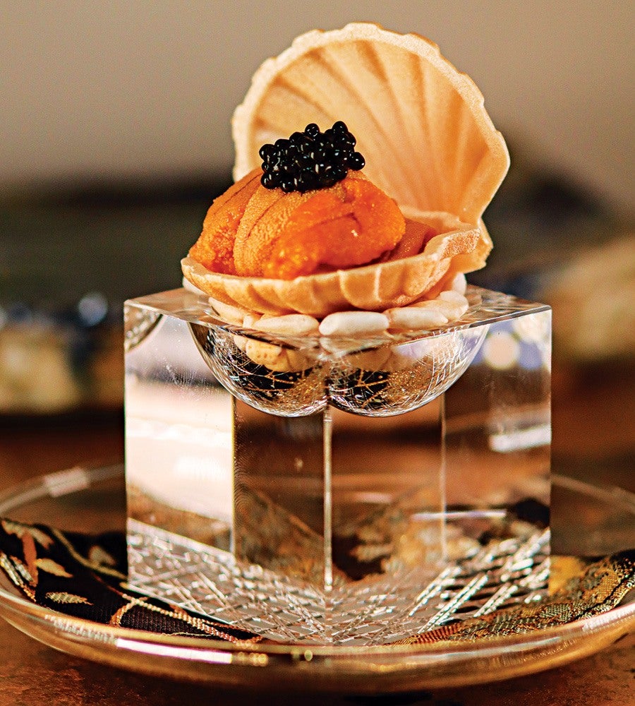 A gourmet dish featuring bright orange sea urchin served in a shell-shaped wafer, topped with a small pile of black caviar. The dish is presented on a clear, faceted glass cube placed on a decorative plate with a patterned cloth underneath.