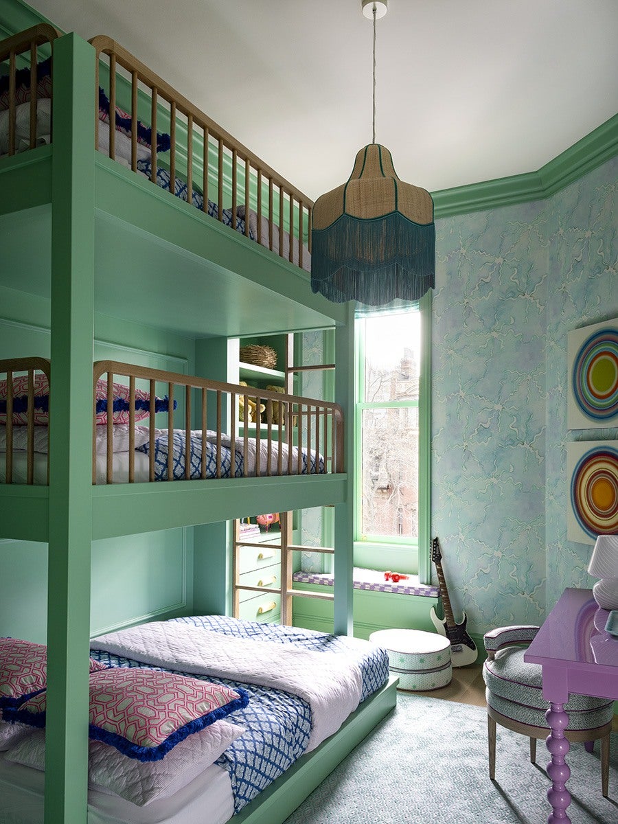 A vibrant bedroom features a mint green triple bunk bed with wooden railings, dressed in blue and pink patterned bedding. The walls are painted with a light blue marbled effect, complemented by mint green trim. A large window with a built-in bench seat lets in natural light, and a black electric guitar rests against the wall. The room includes a pink desk with a matching chair upholstered in a light patterned fabric. A unique pendant light with a beige and teal fringed shade hangs from the ceiling. Two colorful circular abstract paintings are displayed on the wall.