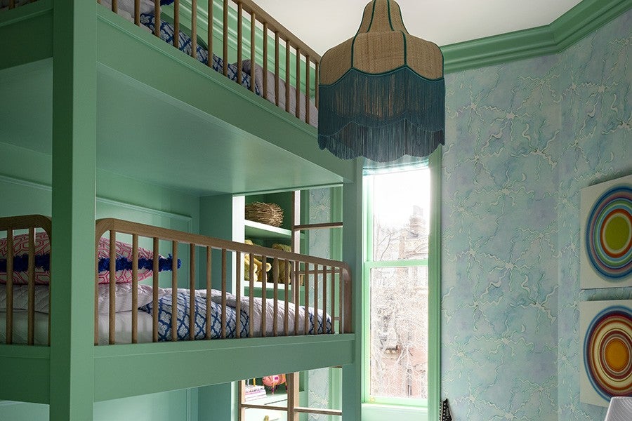 Green-painted wooden bunk beds with wooden railings, featuring blue and pink patterned bedding. A large window with green trim lets in natural light, illuminating a room with light blue marbled wallpaper. A vintage-style hanging lamp with fringe is suspended from the ceiling. Two colorful circular abstract paintings are partially visible on the right wall.