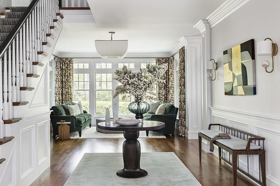 A bright living room with large windows and floral curtains. Two dark green velvet sofas face each other with light-colored pillows. A round wooden table with a large glass vase filled with white flowers is centered on a light rug. To the right, a wooden bench with a cushioned seat and a folded blanket sits against the wall, which features white wainscoting, two wall sconces, and an abstract painting. The floor is wooden, and a staircase with white spindles and wooden handrails is visible on the left.