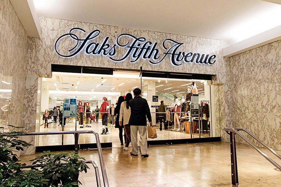 Entrance to a Saks Fifth Avenue store with glass doors, marble walls, and a large illuminated cursive sign above. Several people are walking into the store, and mannequins dressed in fashionable clothing are visible inside.