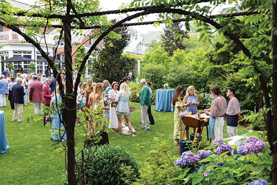 A garden party with a group of people socializing on a well-maintained lawn. The scene is framed by leafy branches and features a mix of men and women dressed in summer attire. There are tables covered with blue tablecloths and a wooden serving station where two men in striped shirts and aprons are attending to guests. Purple hydrangea flowers and lush green bushes add color and vibrancy to the setting. A large brick house with white trim is visible in the background.